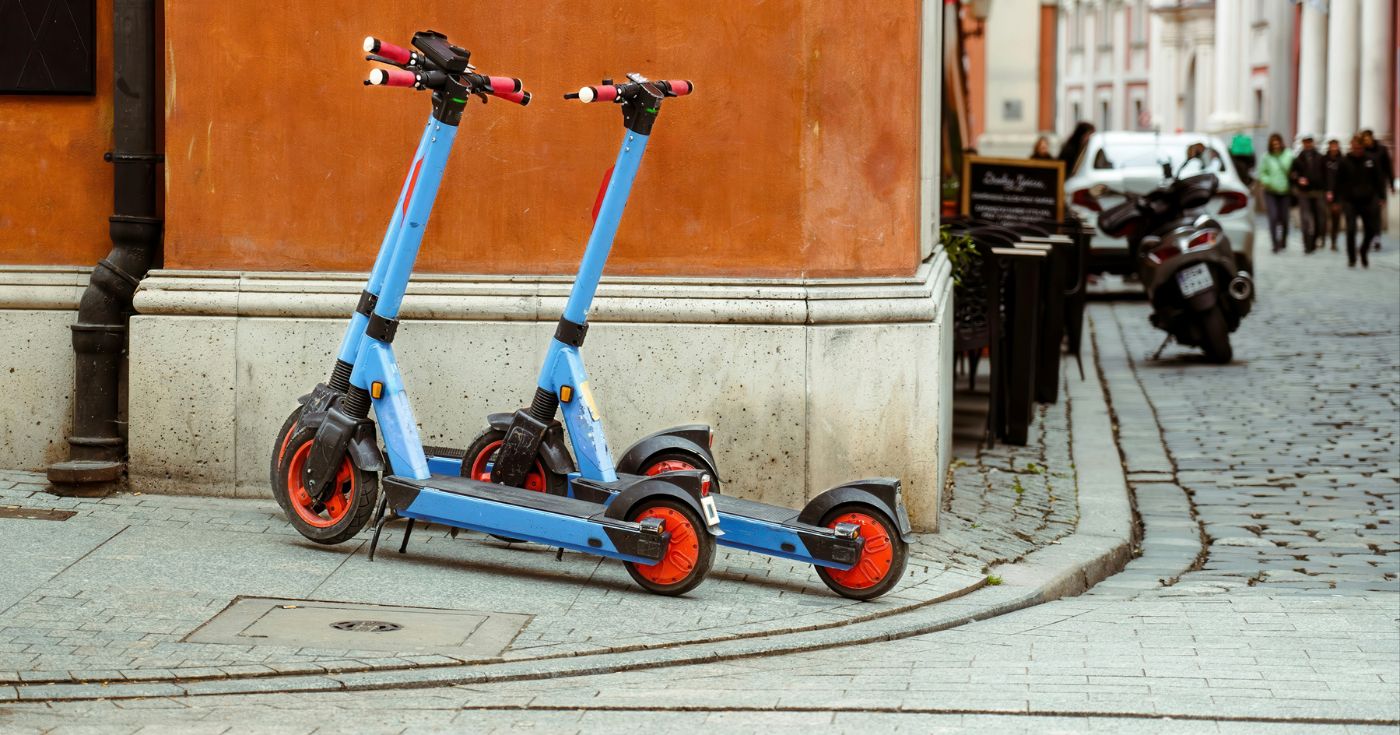 Dwie niebieskie hulajnogi z wypożyczalni zaparkowane pod ścianą 